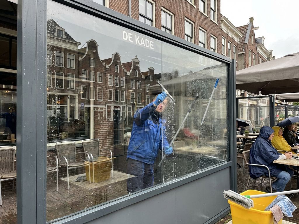 Schoonmaker reinigt glazen windscherm op Nederlands restaurantterras met professionele apparatuur en zichtbare resultaten