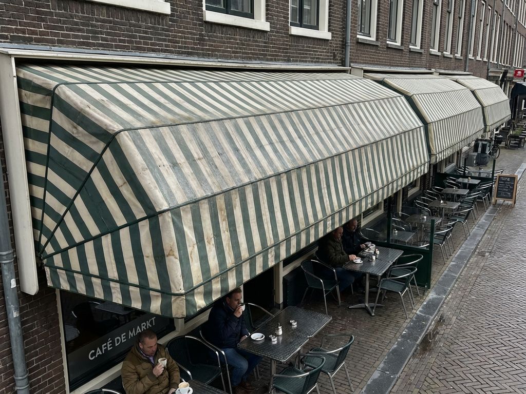 Commercieel terras met gestreepte luifels aan Nederlandse bakstenen gebouw, lichte vervuiling zichtbaar op doek