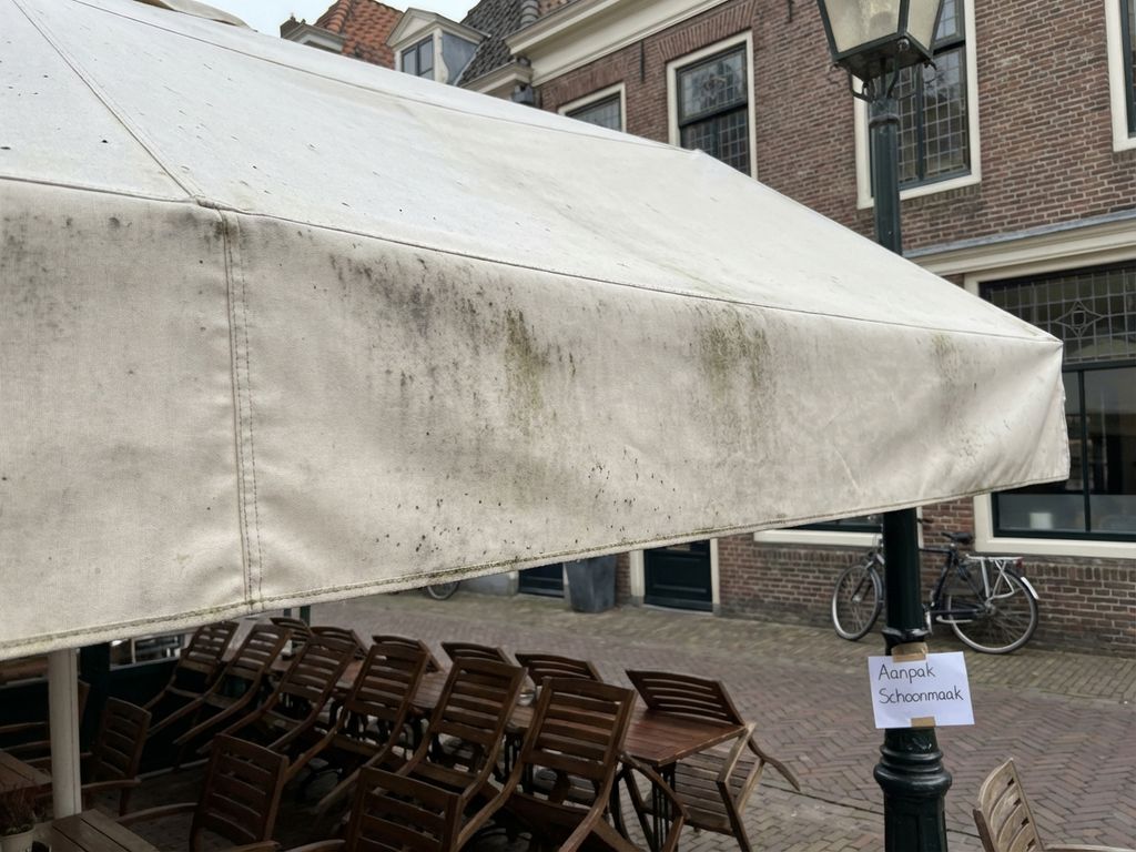 Wit zonnescherm met donkere vlekken en schimmel boven café terras met houten tafels en Nederlandse bakstenen gebouwen