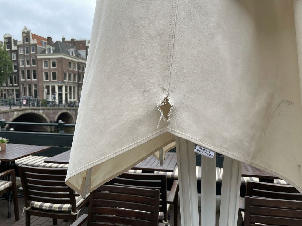 Grote parasol met kleine scheur op Nederlands restaurantterras, houten meubilair en stedelijke architectuur op achtergrond