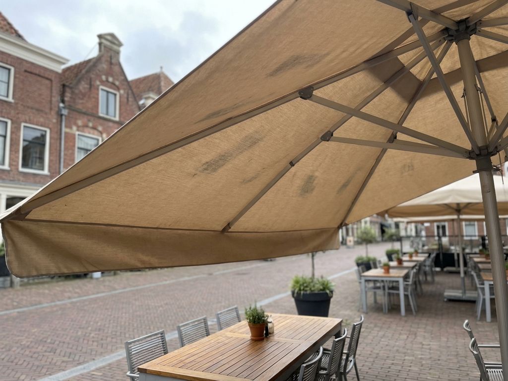 Parasol met beginnende schimmelvorming op beige doek boven restaurantterras met Nederlandse bakstenen gebouwen op achtergrond