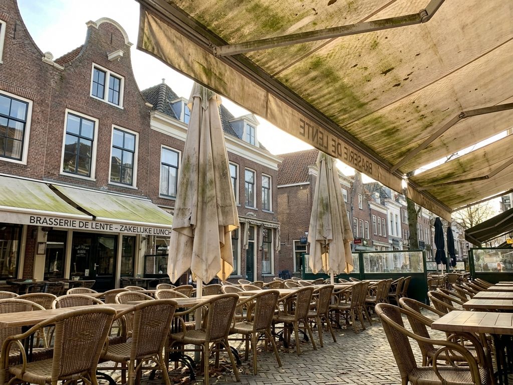 Nederlandse restaurantterras met verweerde zonwering en tuinmeubilair tegen bakstenen gebouwen in voorjaarslicht