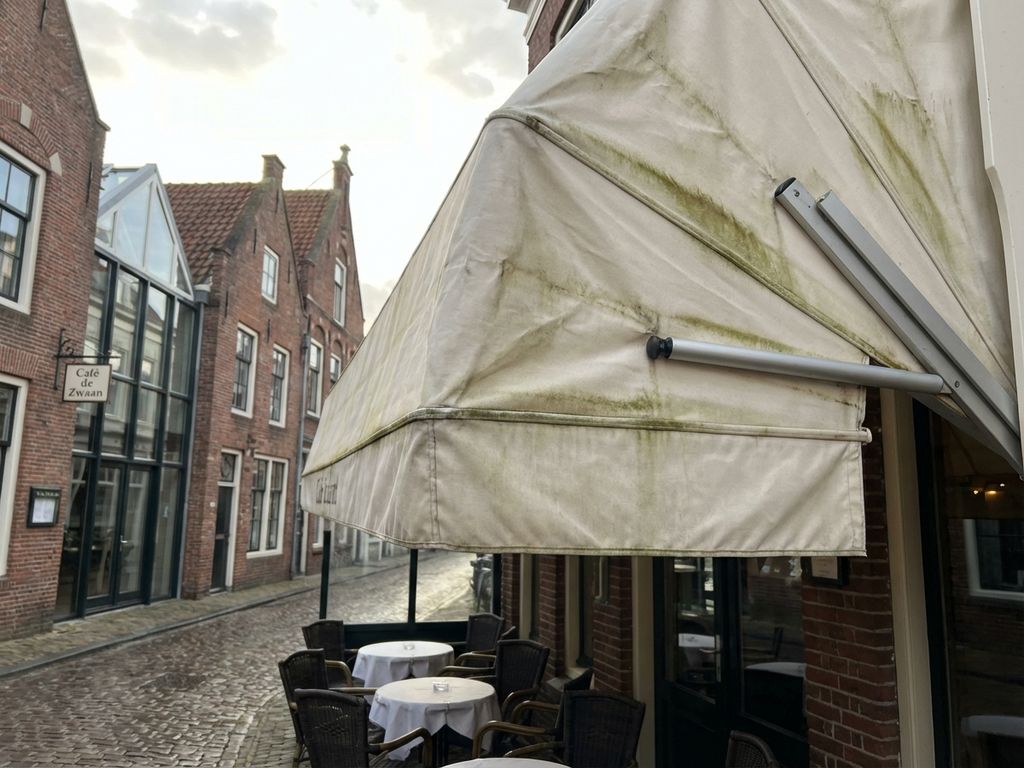 Restaurantterras luifel met lichte algengroei op wit doek, uitgestrekt boven tafels en stoelen in Nederlandse café setting