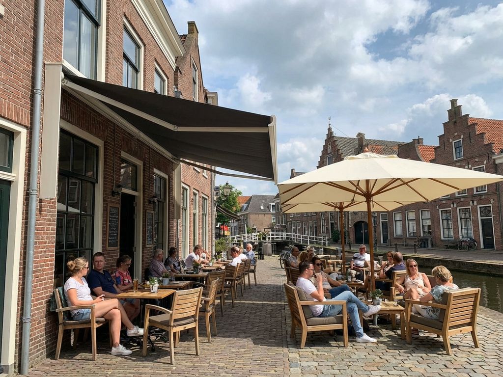 Nederlandse restaurantterras met uitschuifbare zonwering boven tafels en vrijstaande parasol, bakstenen gebouwen op achtergrond