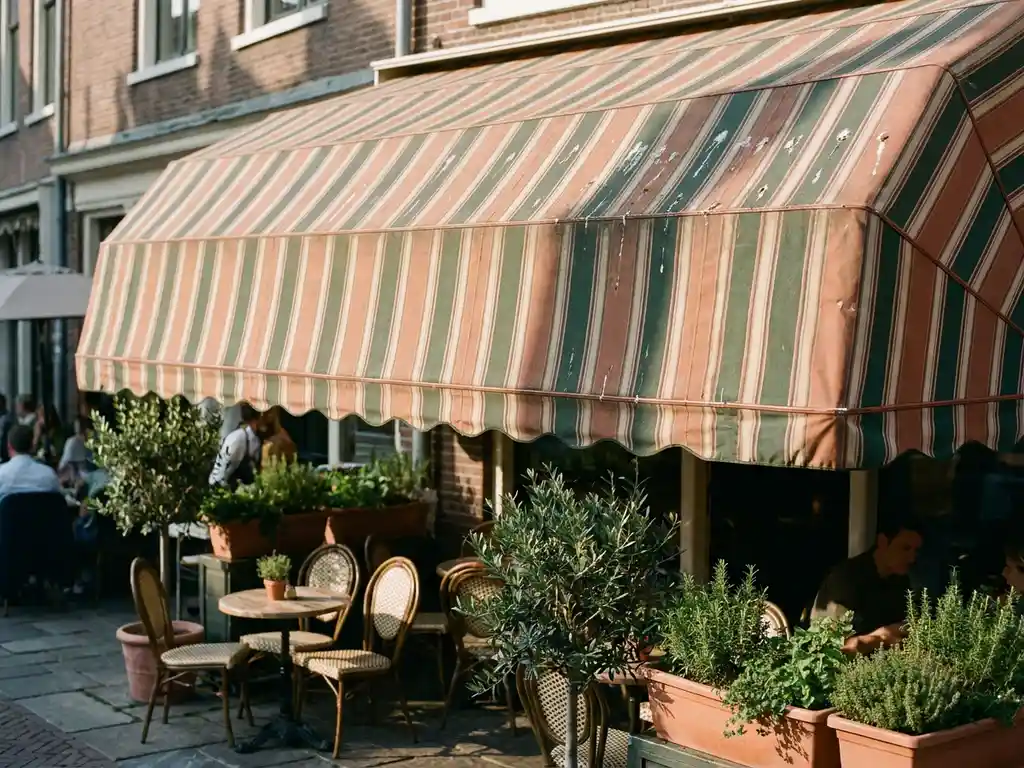 Gestreept canvas terrasscherm boven een Nederlands restaurantterras met vogelpoep, smeedijzeren stoelen en kruiden in potten eronder.