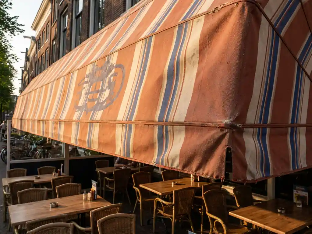 Gestreepte terracotta en kobaltblauwe luifel boven terras van Nederlands restaurant, stoelen en tafels zichtbaar in zacht gouduurlicht.