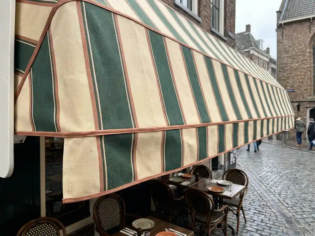 Geïmpregneerd gestreept luifel in crème en bosgroen boven een Nederlands restaurantterras met stoelen, tafels en kasseien op de achtergrond.