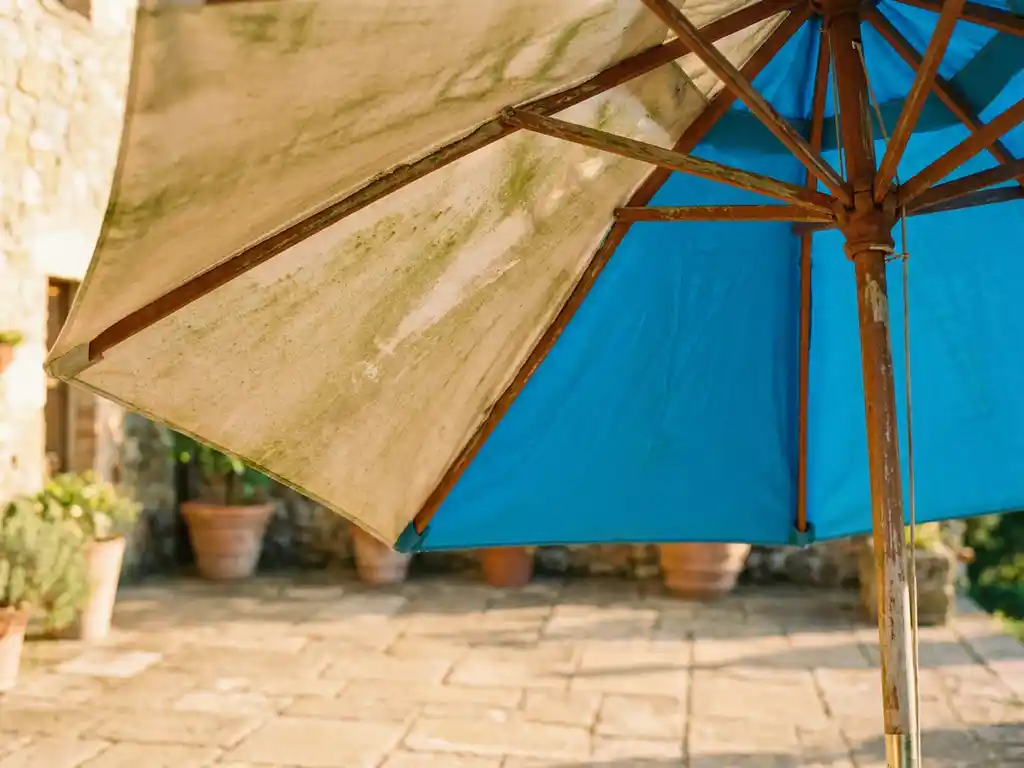 Tuinparasol half verweerd met vervaagde stof en algengroei, half schoongemaakt en kleurhersteld, op een zonnig terras met natuurstenen tegels.
