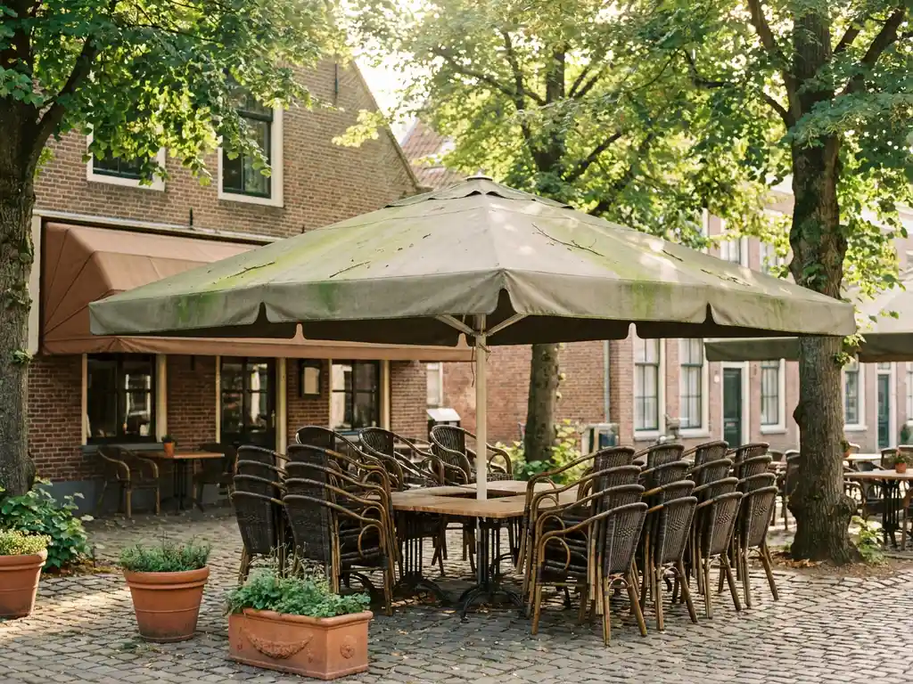 Verweerde parasol met groene algenplekken op een Nederlands restaurantterras met bistrostoelen en kinderkopjes in middagzon.