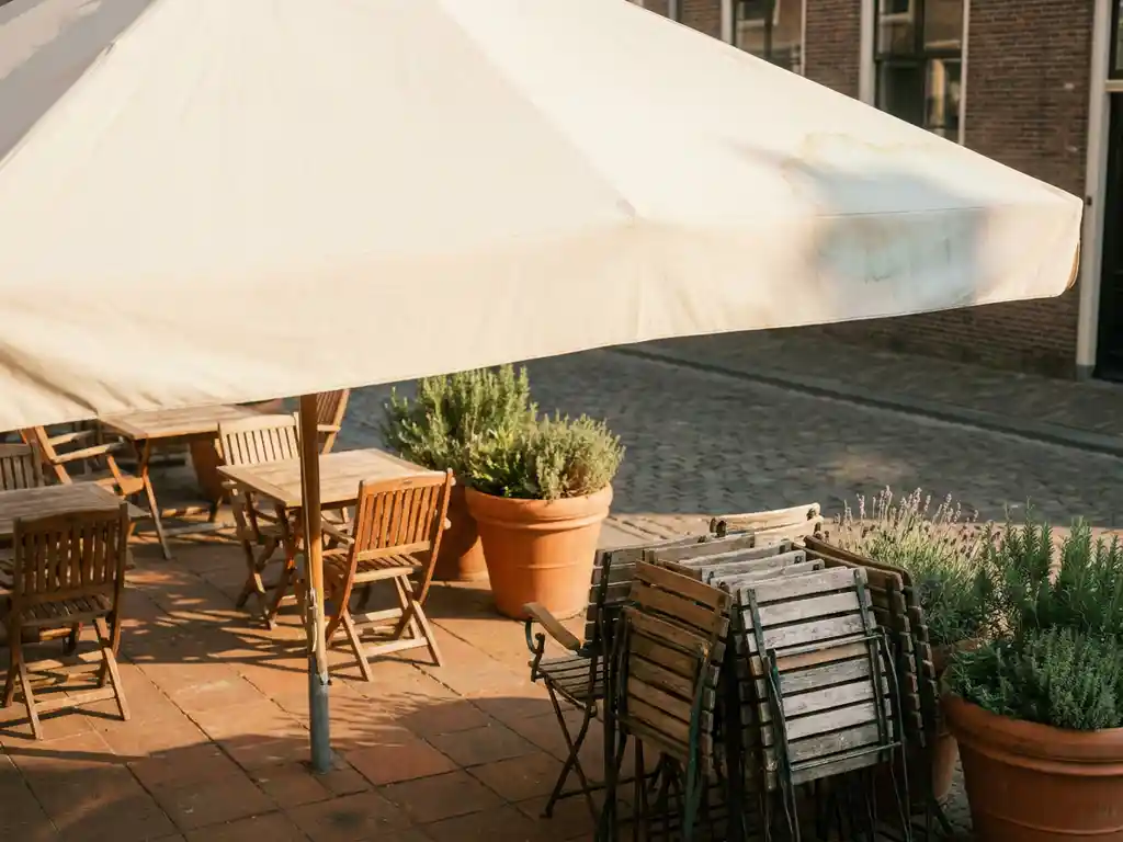 Crèmekleurige parasol op een zonnig Nederlands restaurantterras met houten bistrostoelen, potplanten en terracotta tegels.