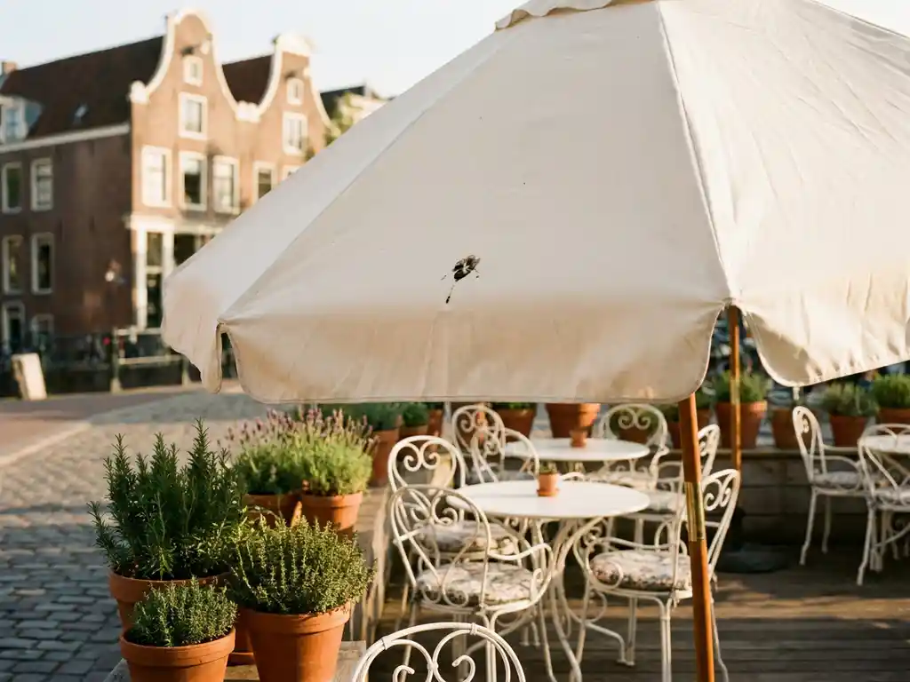 Crèmekleurige parasol op terras van Nederlands restaurant met vogelpoep, witte tuinmeubelen en kruiden, bakstenen gevelachtergrond.