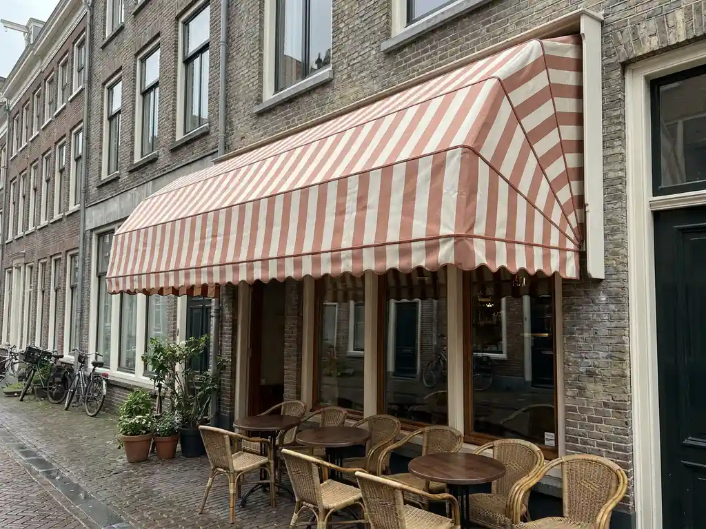 Gereinigd gestreept zonnescherm boven Nederlands café-terras met stoelen en ronde tafels, bakstenen gevel op bewolkte dag.