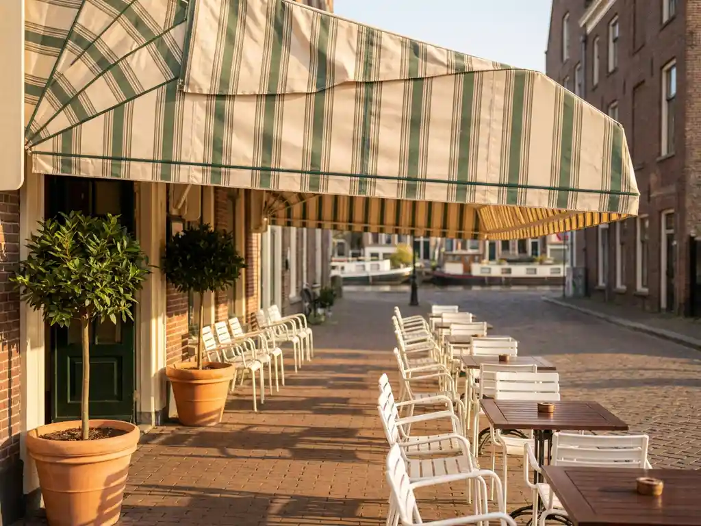 Schoongemaakt gestreept luifel boven terras van Nederlands restaurant met witte bistrostoelen, houten tafels en laurierbomen bij de ingang.