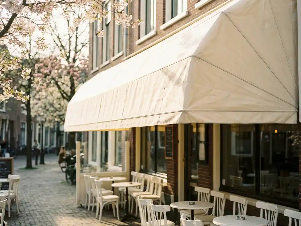 Schoon crèmekleurig restaurantluifel boven een zonnig Nederlands terras met witte bistrostoelen en lentebloesemsem in de ochtendzon.
