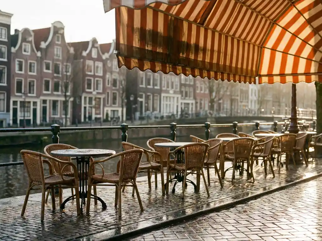 Schoongemaakt terras van Amsterdams restaurant met rieten stoelen en ronde bistrotafels op glanzende natte kinderkopjes, gestreept canvas zonnescherm en grachtenhuizen op achtergrond.