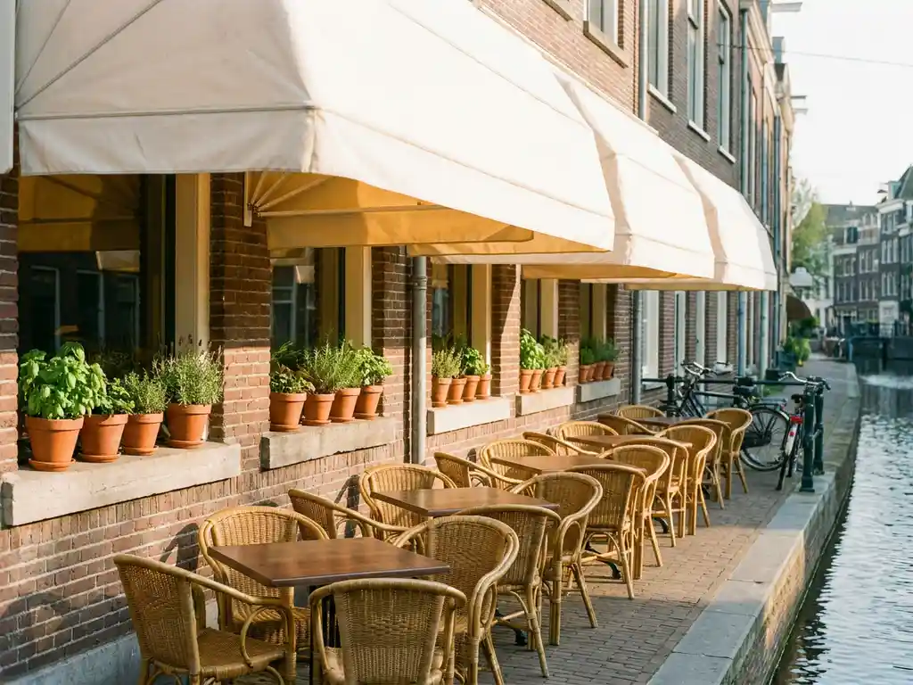 Schoon terras van een restaurantje aan een Amsterdamse gracht met witte zonneschermen, bistrostoelen en kruiden op de vensterbank in ochtendlicht.