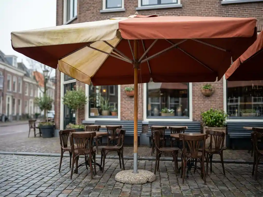 Vervaagde terrasomnbrella met één verkleurde, vergeling paneel boven een Nederlands restaurantterras met houten bistrostoelen en klinkers.