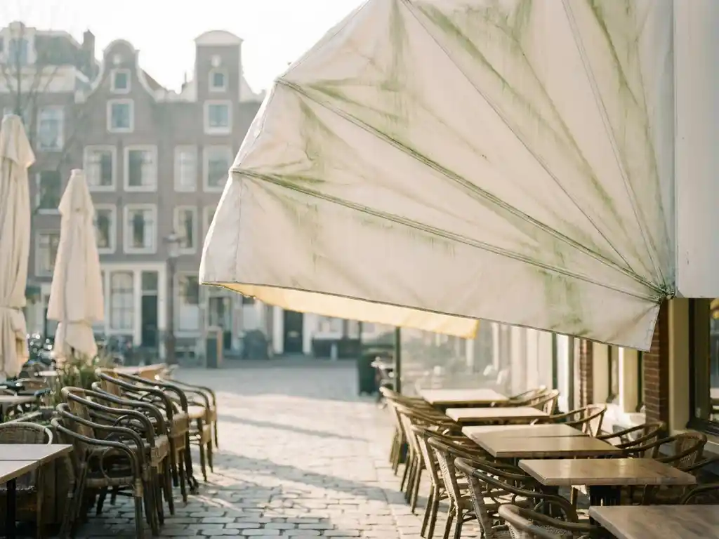 Restaurantterras met crème-wit zonnescherm en groene algenstrepen, lege stoelen bij Hollandse grachtenpanden in vroeg voorjaarslicht.