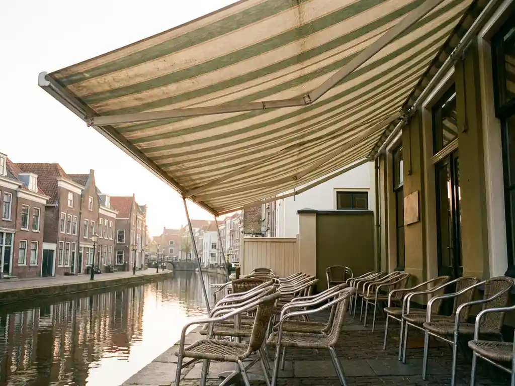 Vervaagd terrasscherm met crème en bosgroene strepen boven een canalside restaurant in Nederland, zacht ochtendlicht op het doek.