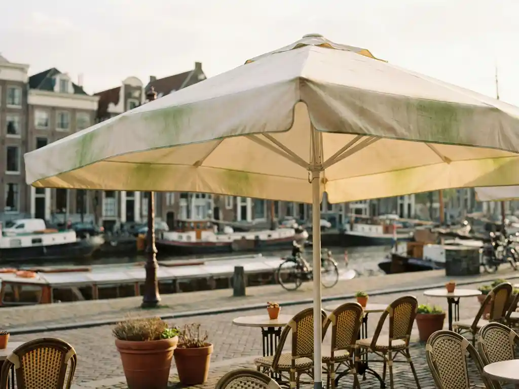 Verweerde terrasparasol op een zonnig Nederlands restaurantterras aan een gracht, met bistrostoelen en kasseien op de achtergrond.