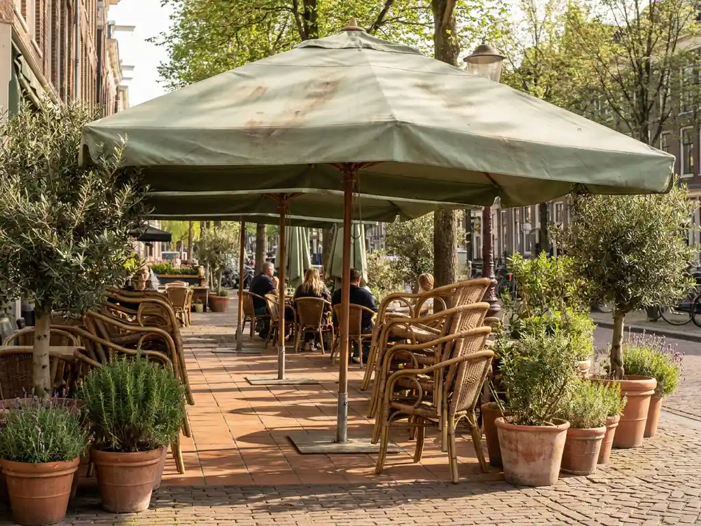 Grote commerciële parasol op een zonnig Nederlands restaurantterras met bistrostoelen, potplanten en terracotta tegels in voorjaarslicht.