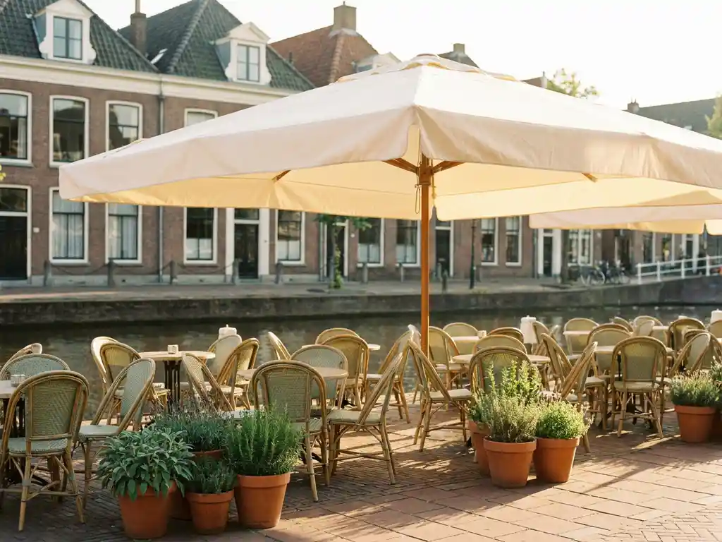 Crème parasol op zonnig terras van Nederlands restaurant aan gracht, met bistrostoelen, potplanten en terracotta tegels in warm middaglicht.