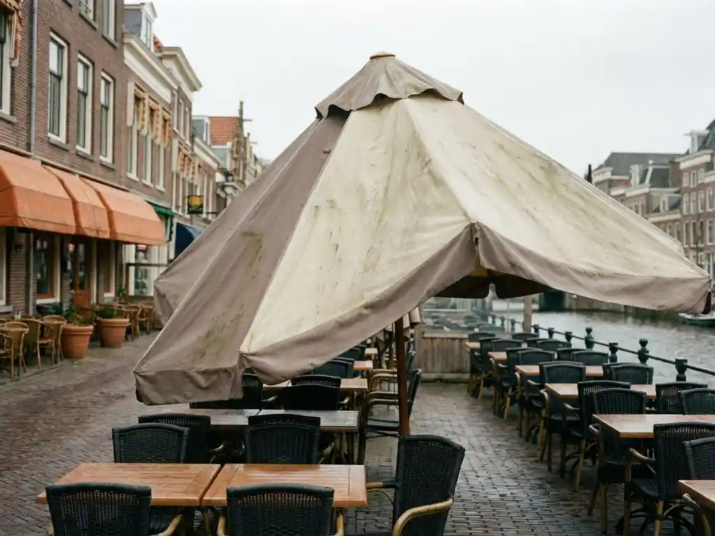 Verweerde terrasparasol met schimmelplekken boven een Nederlands restaurantterras met bistrostoelen en kasseien.