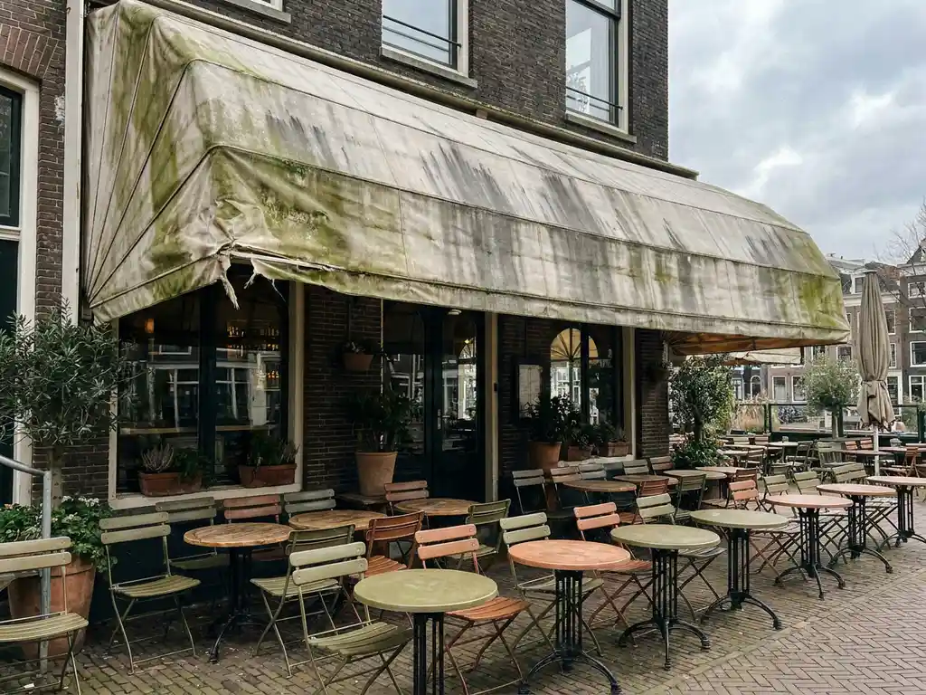Versleten uittrekbare luifel met groene algenplekken boven een Nederlands restaurantterras met bistrostoelen op een klinkerpad.