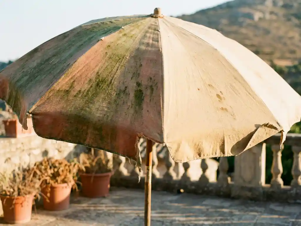 Verweerde parasoldoek met groene algevlekken en rafelige randen naast schoner, helderder stof op zonnig terras.