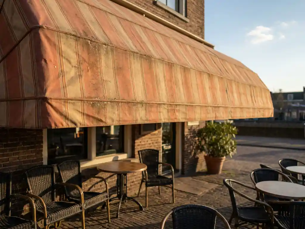 Verbleekt gestreept zonnescherm boven een Nederlands restaurantterras met bistrostoelen op een klinkerbestrating in warm middagzonlicht.