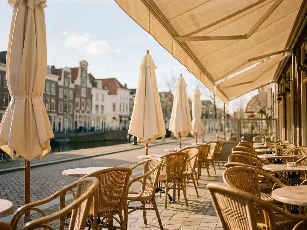 Zonnig terras van een Nederlands restaurant met schone crèmekleurige parasols, rieten stoelen en tegelvloer, grachtenpandarchitectuur op de achtergrond.